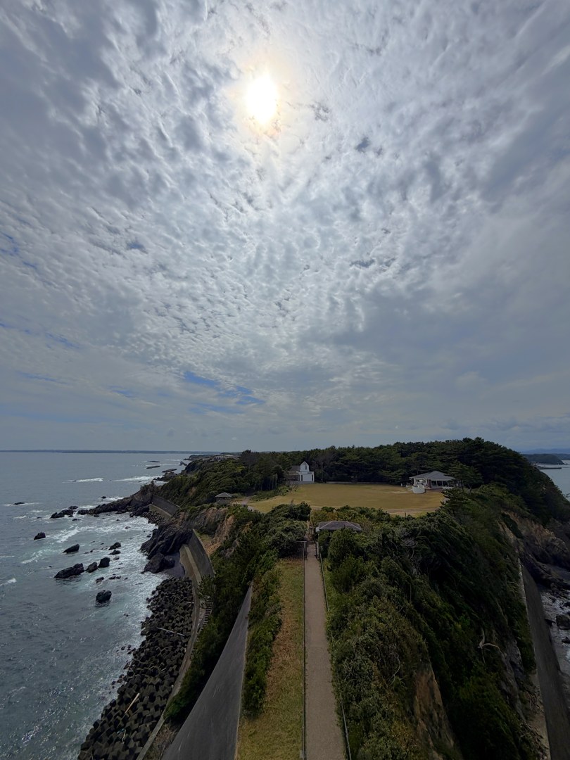 安乗埼灯台の上から見下ろした縦の一枚。上の三分の二は一面のうろこ雲で、左寄りに太陽が雲ごしに白くにじんでいる。下の三分の一は岬。まんなかを細い遊歩道がまっすぐ手前から奥へ伸び、その左に舗装路、さらに左はコンクリートの護岸とテトラポッド、そして岩に白波の立つ海。道の両側は緑の茂み。奥に芝生の広場と、屋根の見える小さな建物がいくつか。