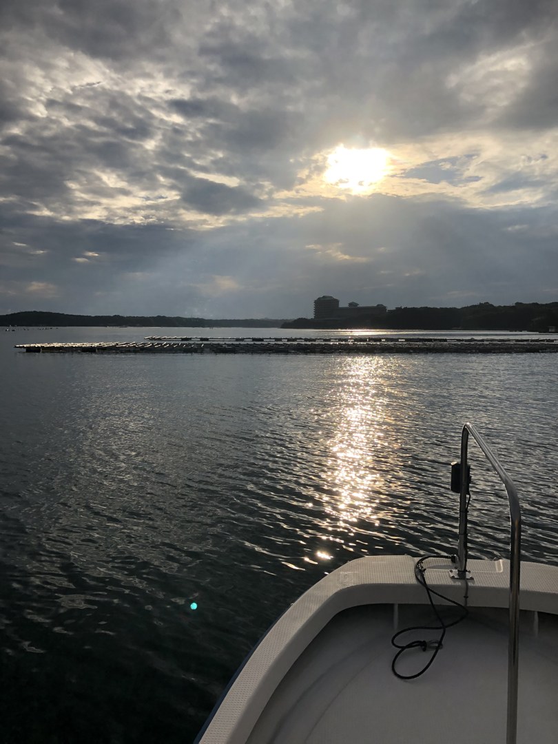 夕方の海を、船の上から撮った一枚。空の上半分は厚い灰色の雲におおわれ、その切れ間から白い太陽がのぞいている。海の真ん中に、太陽の光が縦一本の帯になって手前まで伸びている。左から右へ、真珠いかだの列が水平線のように連なり、その向こうに対岸の陸地と建物が小さく見える。画面の右下に、白い船べりと細い手すり、黒いコードが写り込んでいる。