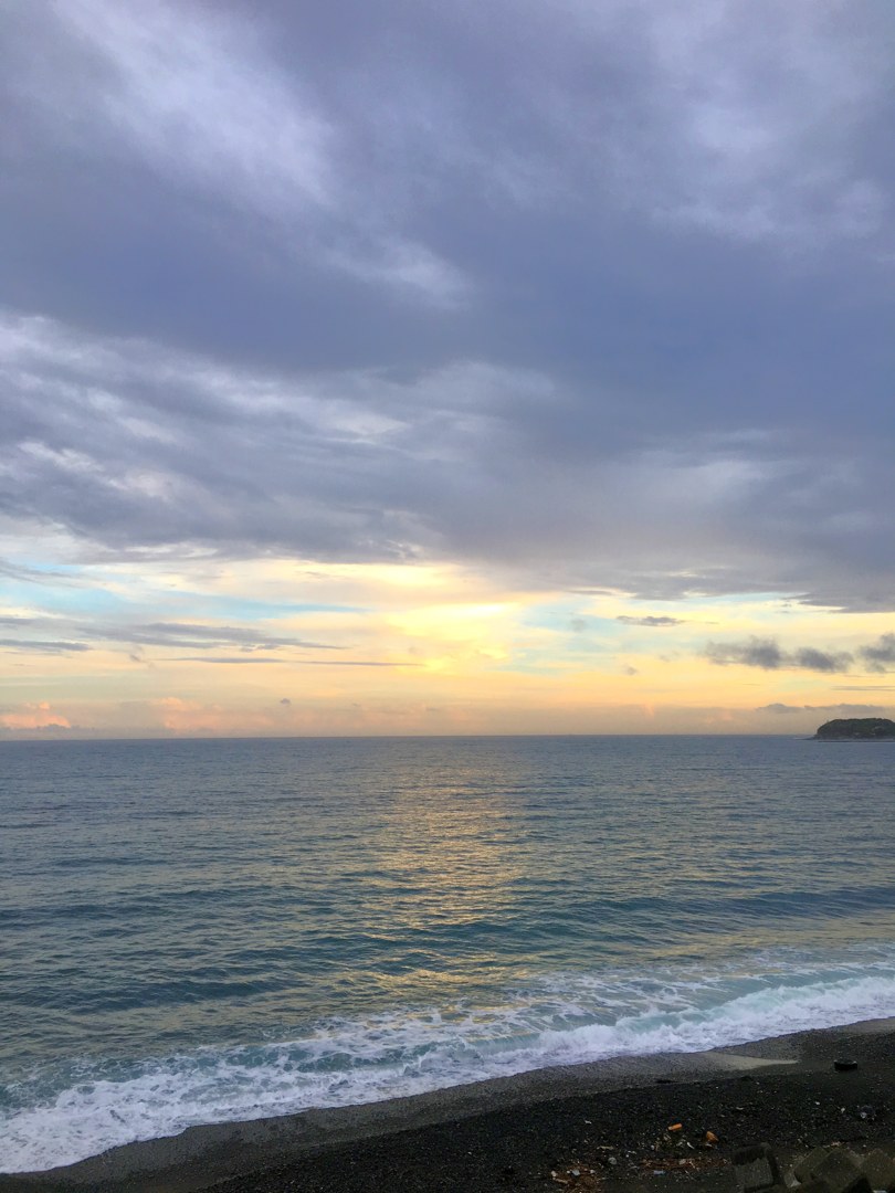 ぶあつい雲が垂れた夕方のひろの浜。水平線のすぐ上だけが金色に空いていて、手前の黒い砂利の浜に波が斜めに寄せている。右奥に岬。
