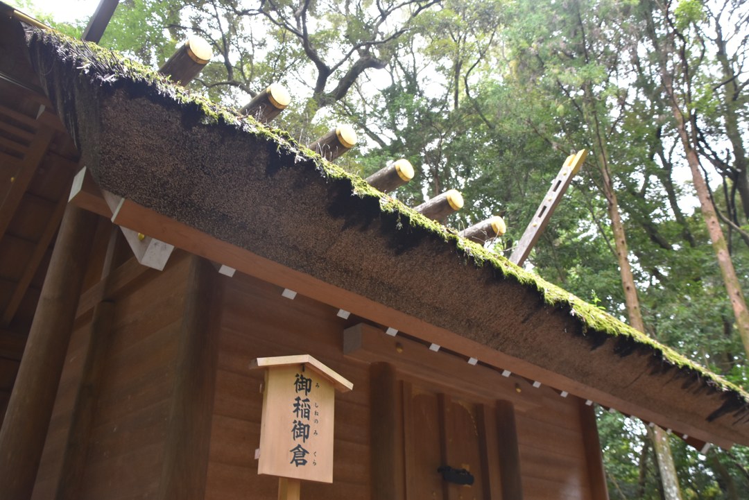 木立のなかの小さなお社を見上げたところ。茅葺きの屋根の上に苔が緑にのっていて、棟には白木の千木と鰹木が並んでいる。手前の柱に『御稲御倉』と書かれた白木の札。