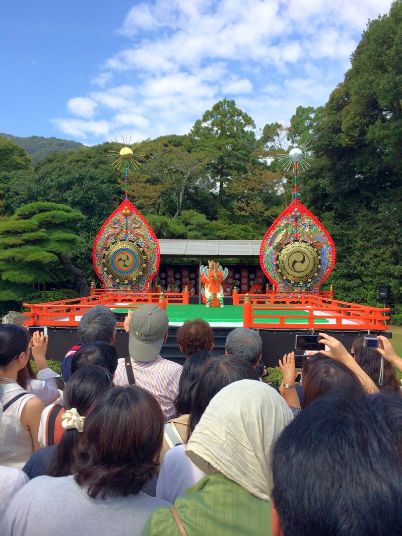 内宮の神苑の特設舞台。左右に大きな赤い飾りの太鼓が立ち、まん中でオレンジの装束の舞い手が舞っている。手前にはたくさんの人の後ろ姿があり、何人かがスマホを高くかざしている。空は青く、白い雲。