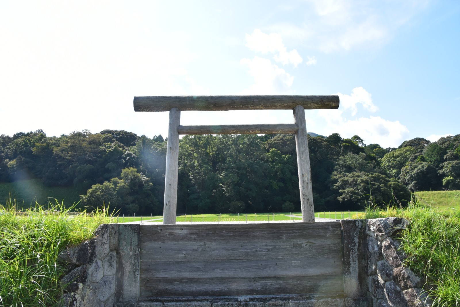 石段と木の板塀の上に立つ、伊勢神宮式の白木の鳥居。柱のあいだから、みどりの田んぼと、その奥の深い森が見える。空は明るく、白い雲がうかんでいる