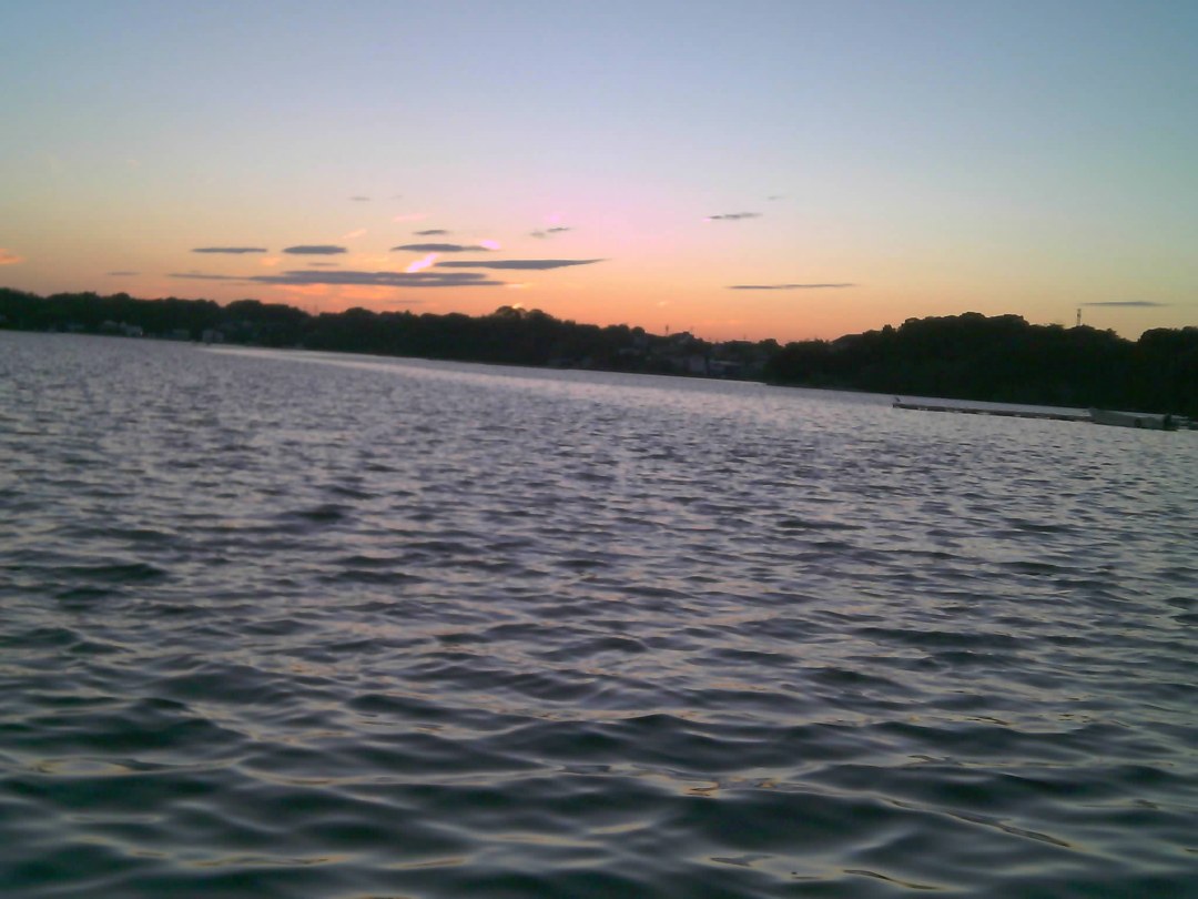 夕暮れの海。画面の下三分の二が細かく波立つ水面で、残りが空。対岸は木立の黒いシルエットになり、空は下からオレンジ、ピンク、水色へと移っていく。細い雲が横に数本