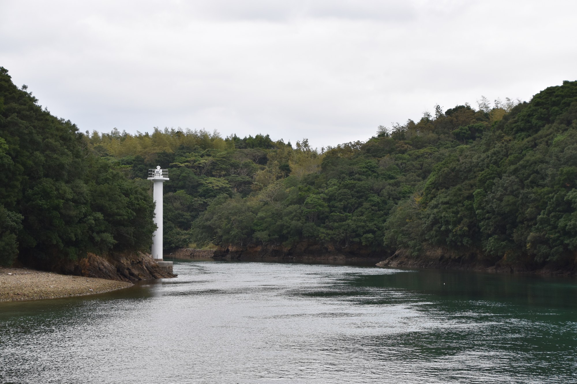 志摩市の深谷水道。曇り空の下、緑の山にはさまれた入り江の口に白い灯台がひとつ立ち、深い青緑の水がゆるやかに流れている