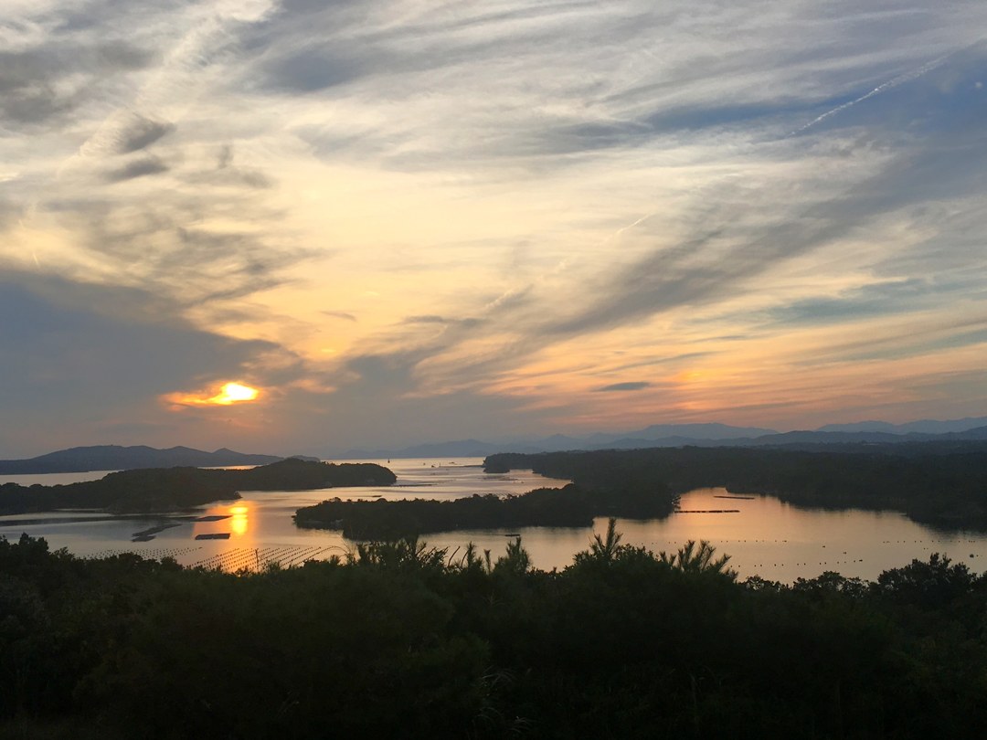 高い場所から見下ろす入り江と島々。沈みかけた夕日が水面に一本の光の筋を落としている