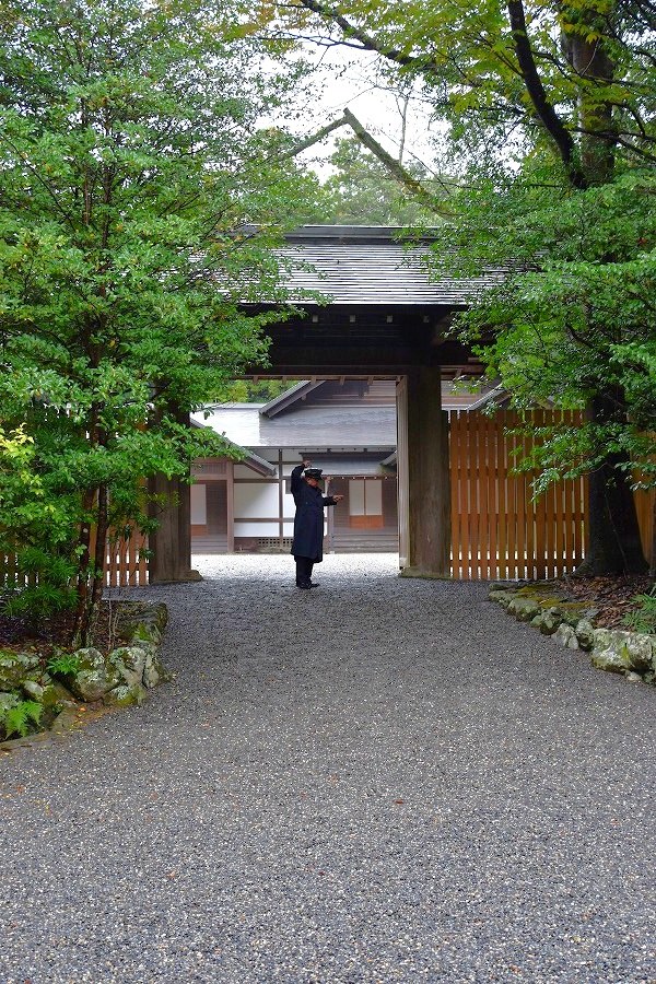 砂利の参道の先に素木の門。門の奥に人がひとり小さく立っている
