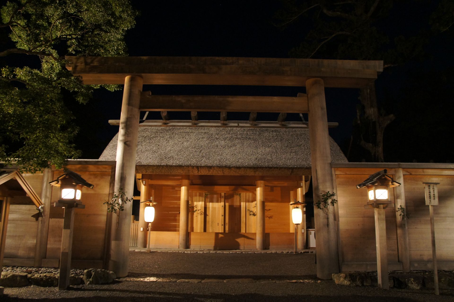 夜の正宮の門。灯りがともり、茅葺きの屋根の下の扉が橙色に照らされている