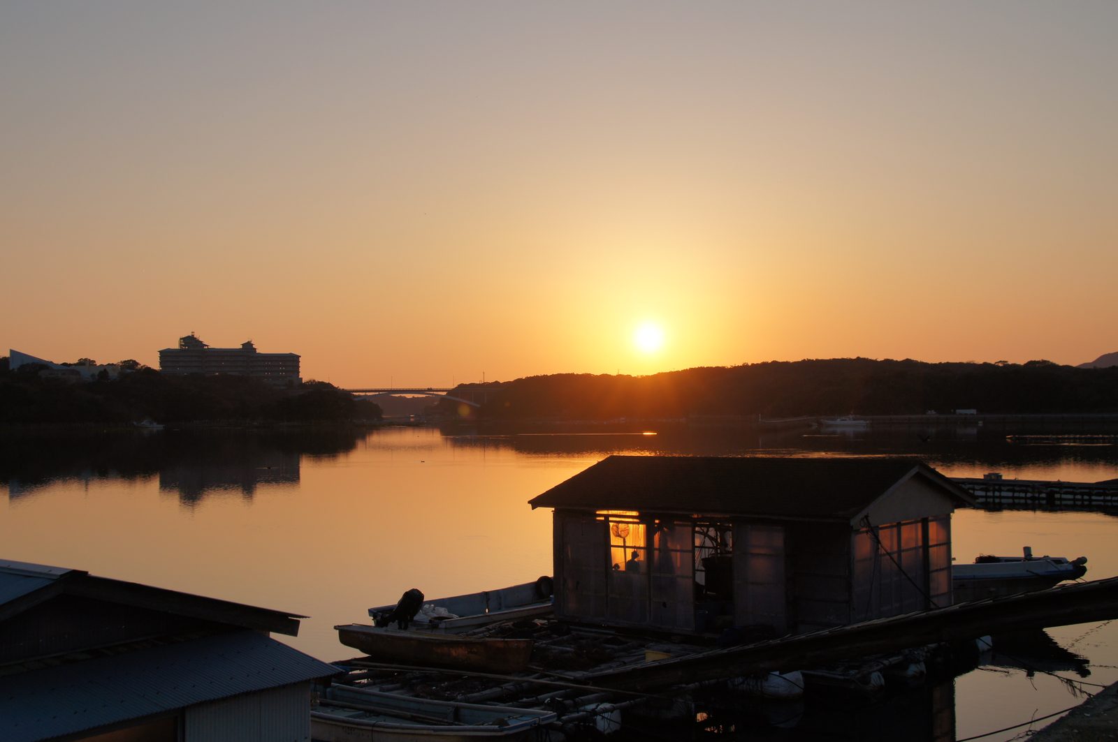 賢島大橋のあたりの夕日。手前に真珠養殖の作業小屋と小舟、その向こうの丘へ日が沈んでいく
